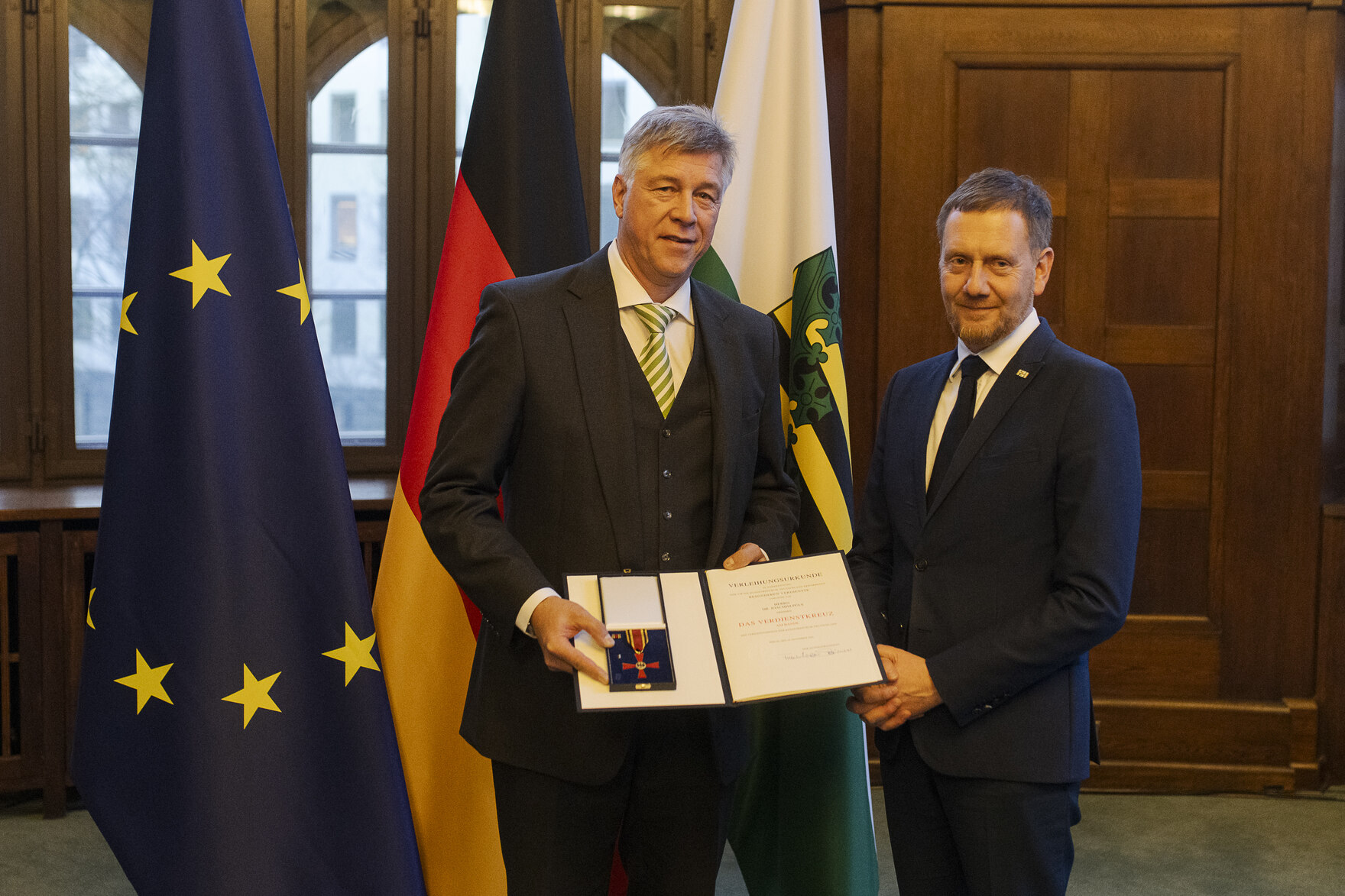 Zwei Männer in einem Raum, der Mann links hält eine offene Präsentationsmappe mit einer Auszeichnung, im Hintergrund die Flaggen der EU, Deutschlands und Sachsens.